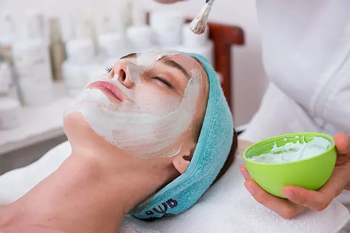 Esthetician brushing a smooth white facial mask onto a relaxed client's face